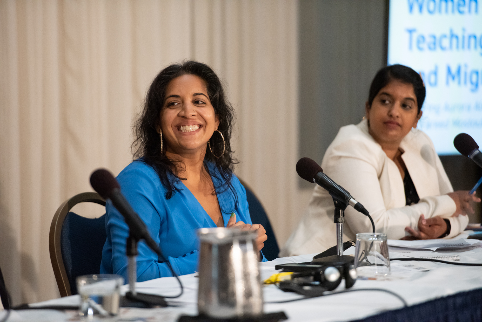 A woman sitting at a table with a microphone smiles with another woman sitting next to her