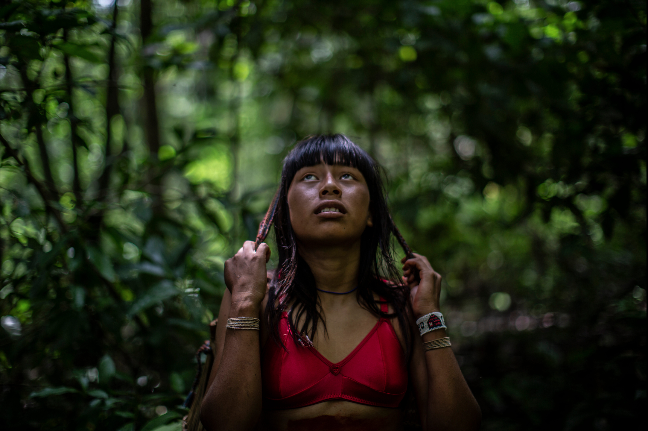 A woman clasps sections of her hair in her hands and gazes up earnestly into the rainforest foliage.